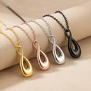 Four necklaces with teardrop pendants in gold, rose gold, silver, and black on a beige surface.