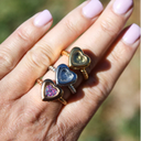 Hand wearing three heart-shaped rings with colorful stones on a blurred natural background